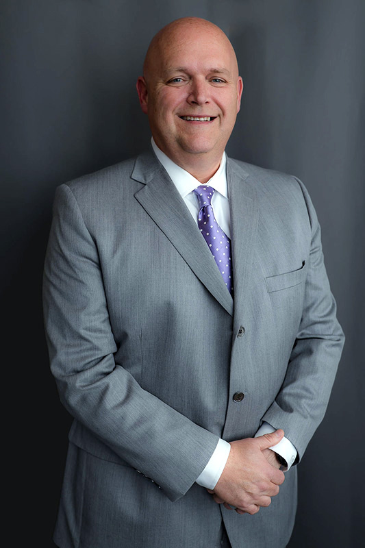 kevin-shearer-county-attorney Kevin S. Shearer in a gray suit and purple tie, smiling in front of a dark gray background, professional portrait for Russell County Attorney office.