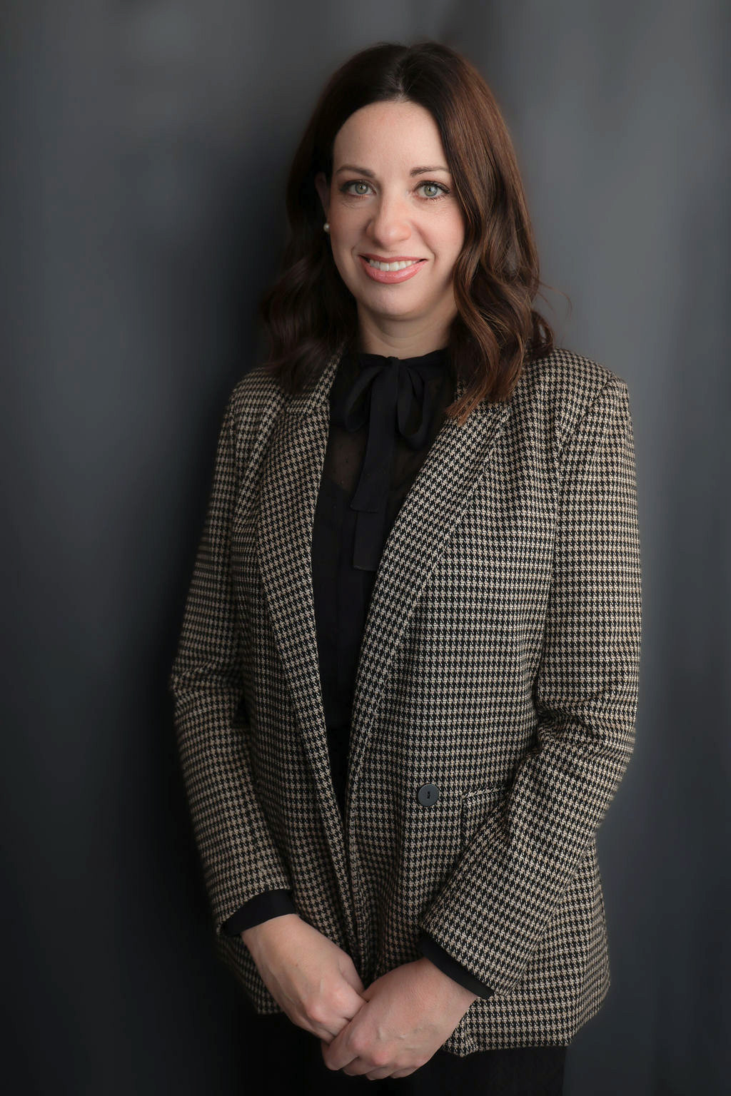 Professional headshot of a woman in a houndstooth blazer and black blouse, smiling in front of a dark gray background.