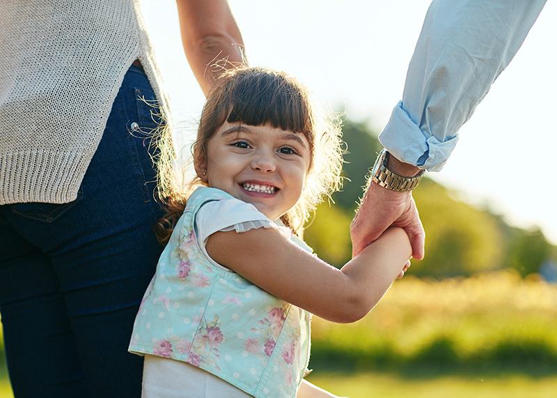 supporting-children-families Young girl smiling and holding hands with adults, symbolizing child safety, family support, and guardianship in a loving outdoor setting.