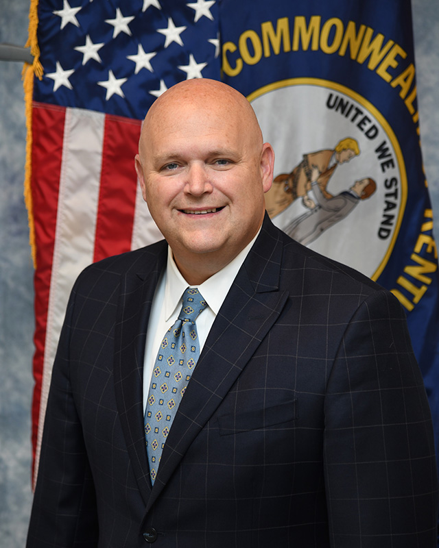 kevin-shearer-russell-county-attorney Kevin S. Shearer, Russell County Attorney, in a suit and tie standing in front of the U.S. and Kentucky state flags.